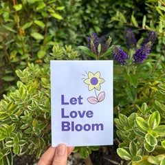 A person holding our "Let love bloom" seed packet in front of green bushes.