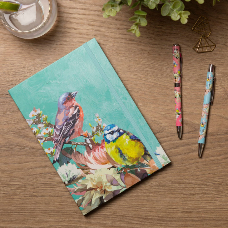 A hardcover notebook with a colorful bird illustration on the cover, placed on a wooden tabletop next to two floral pens and a small glass cup.
